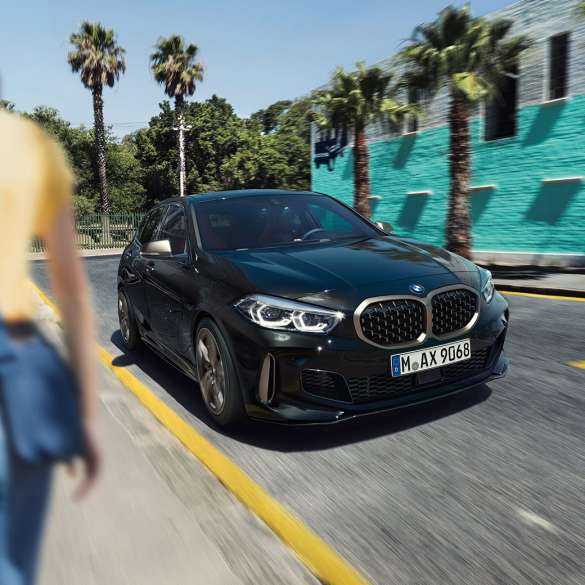 BMW M135i xDrive, vue de trois quarts avant. BMW M135i xDrive en Saphirschwarz metallic, vue de trois quarts avant, avec femme à l’avant-plan.