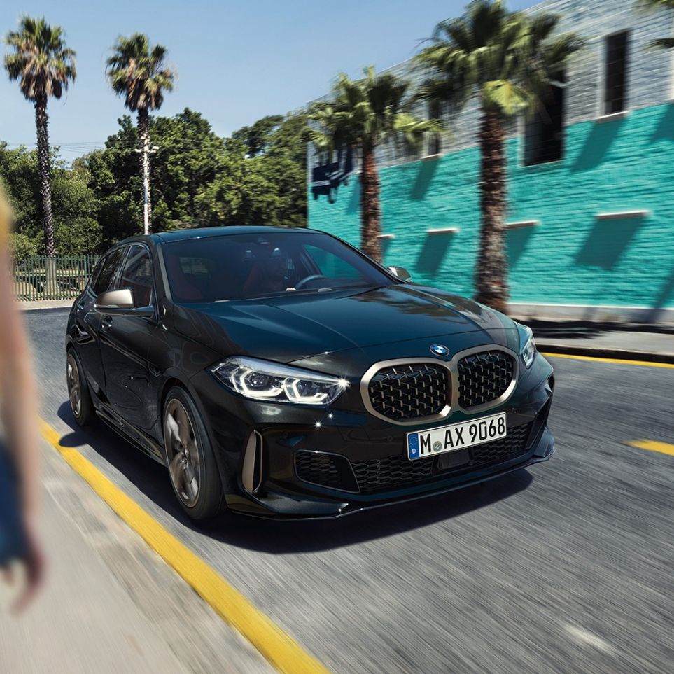 BMW M135i xDrive, vue de trois quarts avant. BMW M135i xDrive en Saphirschwarz metallic, vue de trois quarts avant, avec femme à l’avant-plan.