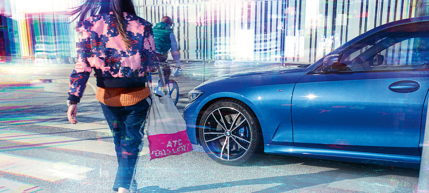 BMW Série 3 Berline, vue latérale de trois quarts. Vue latérale de trois quarts de la BMW Série 3 Berline sur l’asphalte devant un décor urbain avec passants.
