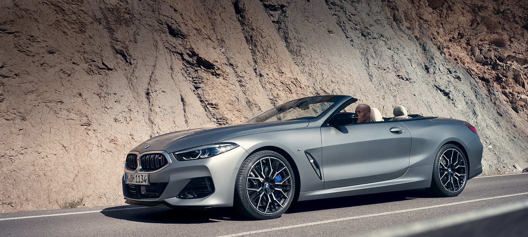BMW 8 Series Convertible G14 LCI Facelift 2022 with open softtop driving along mountain wall BMW 8 Series Convertible G14 LCI Facelift 2022 BMW Individual Frozen Pure Grey metallic side view with open softtop driving along mountain wall