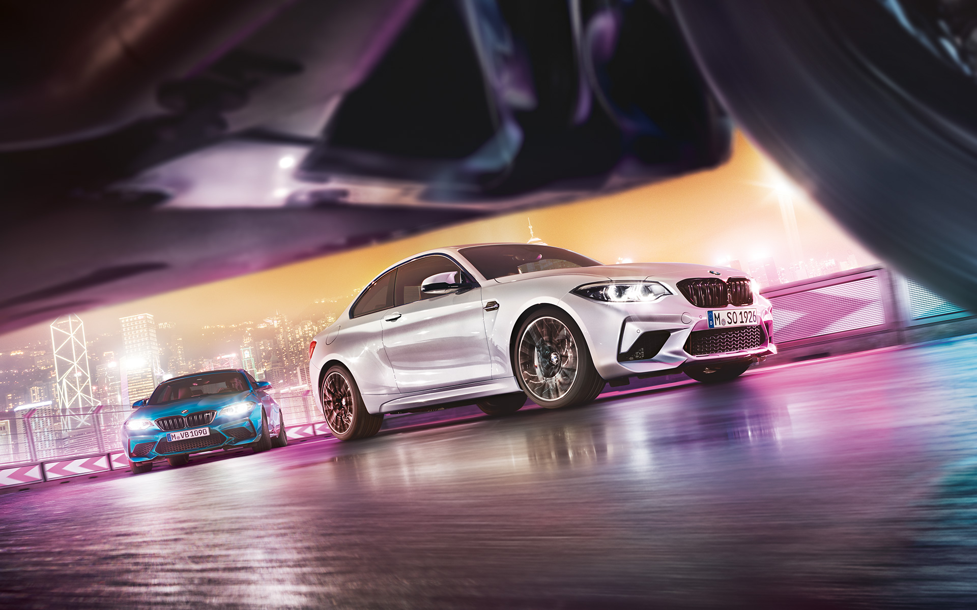 BMW M2 Competition sur du bitume réfléchissant Deux BMW M2 Competition sur le circuit de course, dans le décor nocturne d'une grande ville, vue depuis le sol.