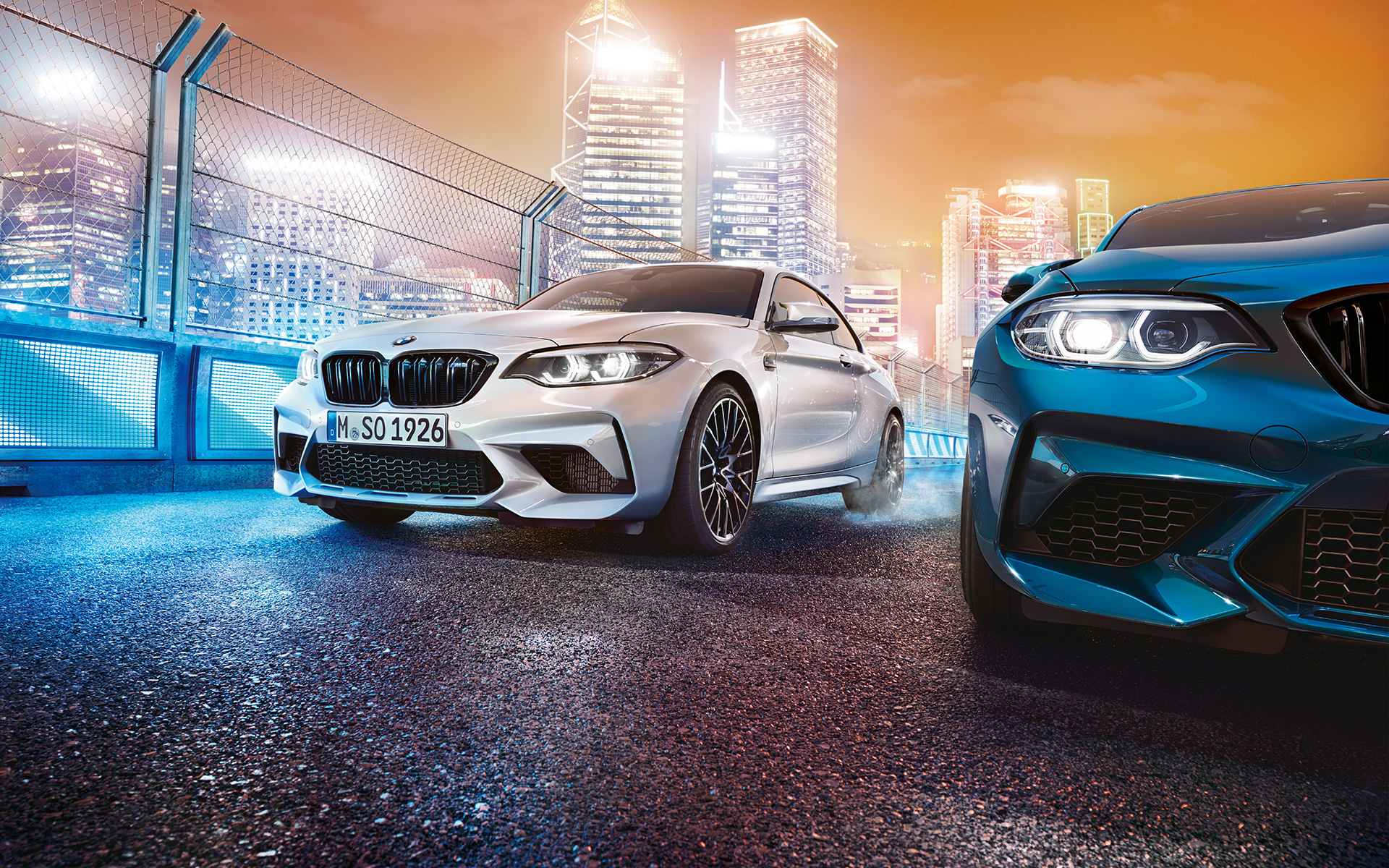 Des BMW M2 Competition blanche et bleue à l'arrêt. Une BMW M2 Competition blanche et une autre bleue vues de l'avant, dans le décor nocturne d'une grande ville.