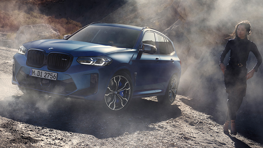 BMW X3 M Competition F97 LCI 2021 vue de trois quarts avant en stationnement sur un terrain rocailleux BMW X3 M Competition F97 LCI restylage 2021 Frozen Marina Bay Blau metallic vue de trois quarts avant en stationnement sur un terrain rocailleux avec mannequin femme