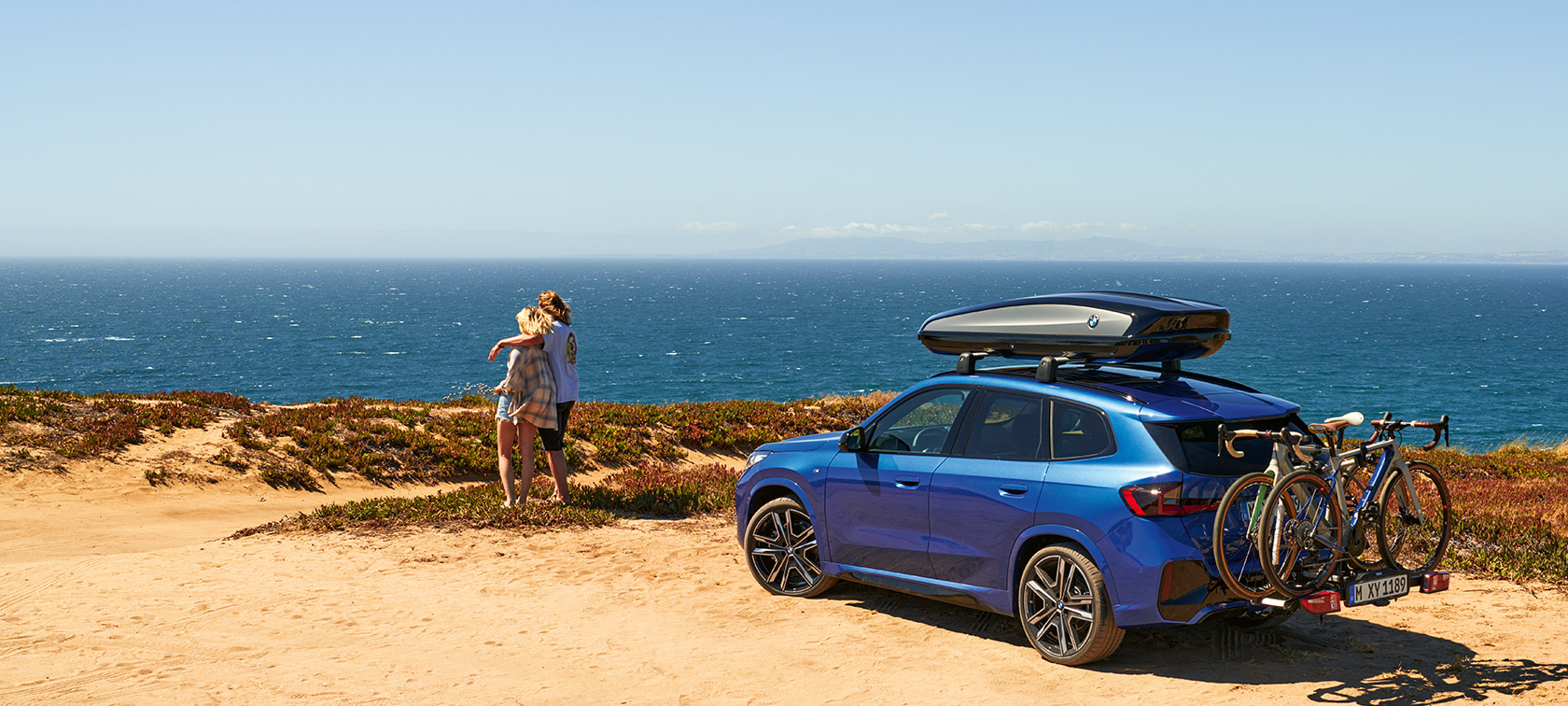 Accessoires d'origine BMW pour le printemps et l’été BMW X1 U11 Accessoires d'origine BMW pour printemps et été BMW X1 U11 à l’arrêt sur le sable devant une côte avec 2 personnes